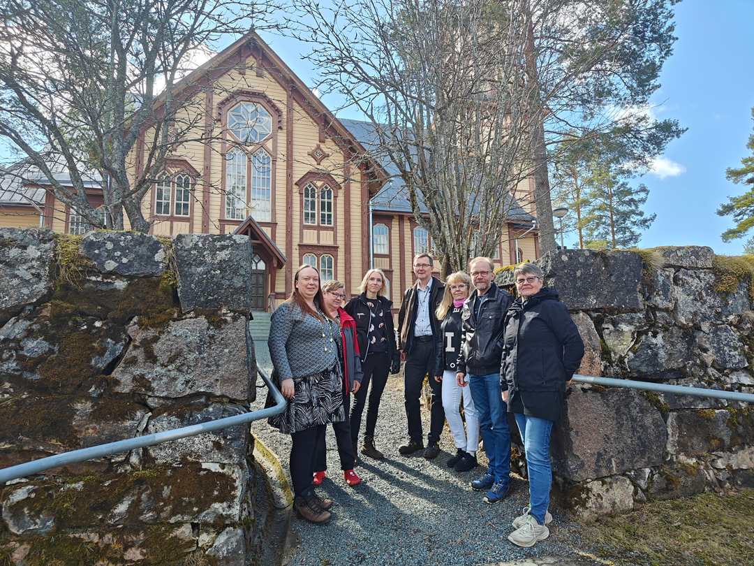 Seitsemän henkilöä Lapinlahden kirkon edustalla kiviaidan juuressa katsovat kameraan. Kirkko taustalla.