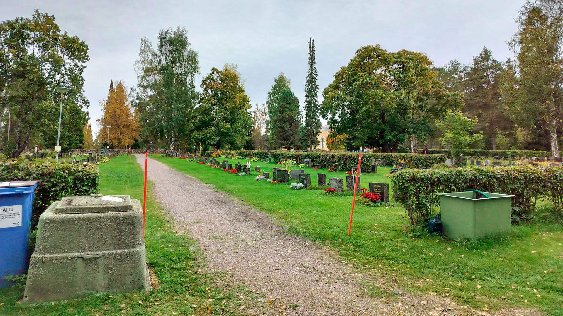 Hautausmaan hiekkakäytävä, hautakivia ja nurmikkoa, jäteastioita.